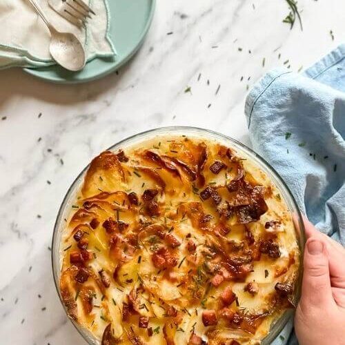 air fryer scalloped potatoes in a tray, under a blue towel, on the side a plate with a towel and cutlery, under a marble background, hand holding the tray