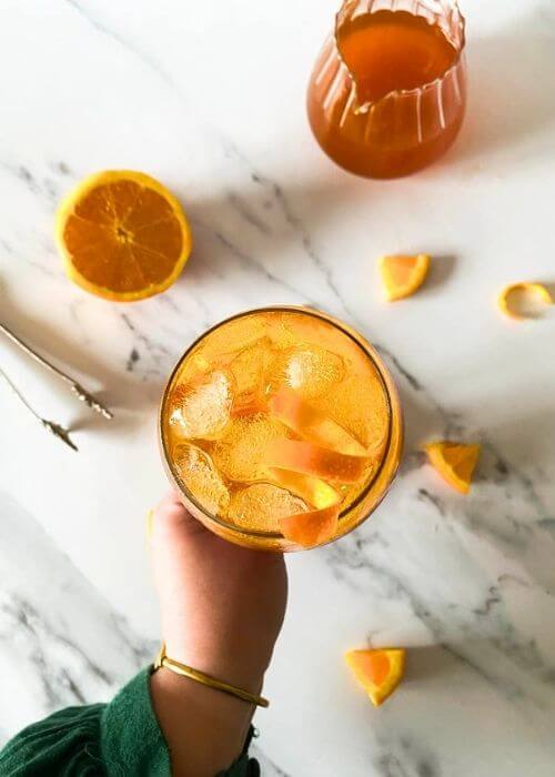 Super Aperol Spritz Mocktail, Just Like The Real One 3 from top - hand holding aperol spritz mocktail, marble background, ice tongues and slices of orange around