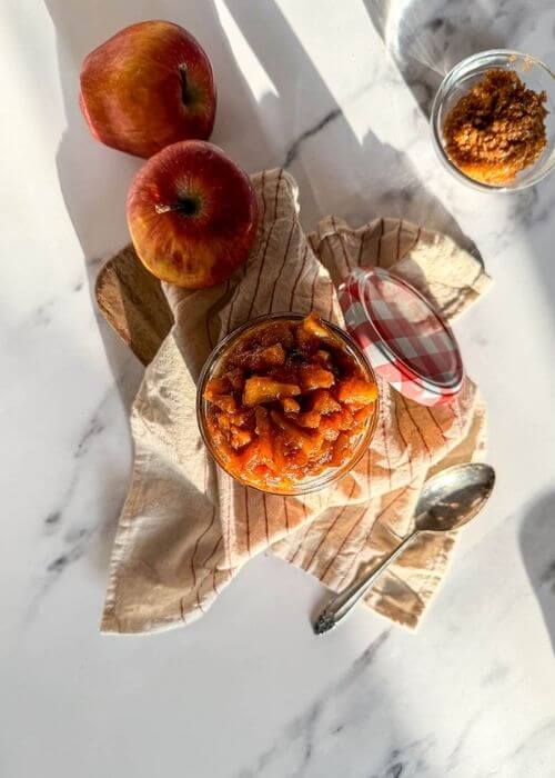 apples caramelized in a jar, over a striped towel, a spoon, apples and brown sugar on the sides, over a marble background