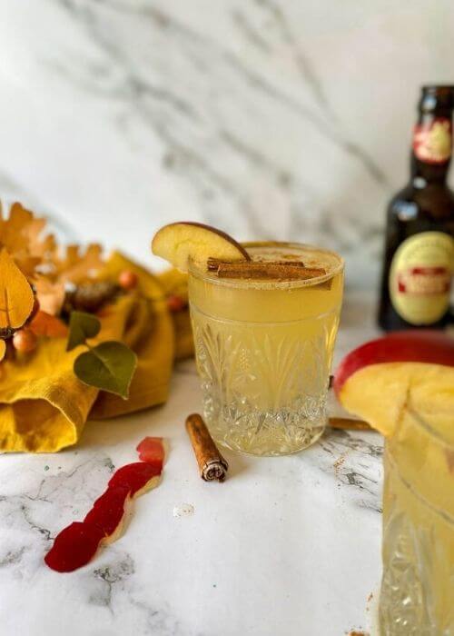 Quick Apple Fizz Mocktail, Delicious Fall Drink 6 two glasses of apple fizz mocktail with apple slices and cinnamon sticks on top, apple peels, cinnamon sticks, knife, towel and leaves around, on marble background