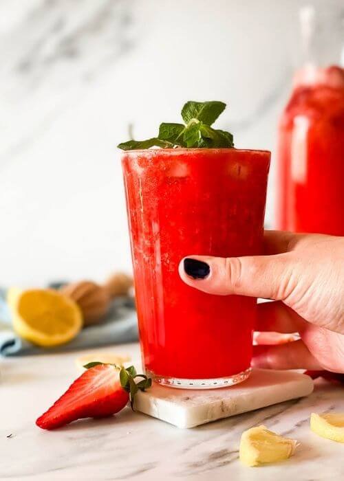 hand holding glass of blended strawberry lemonade on a marble coaster, behind a jug of lemonade, strawberries mint and lemons around, in the back a blue towel and lemon squeezer on a marble background