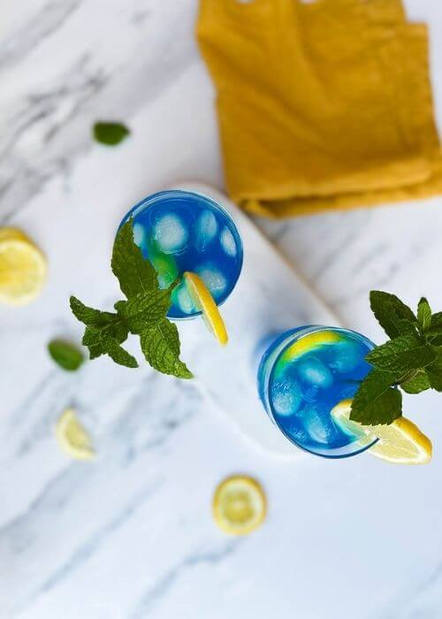 from top - two glasses of blue lagoon fizz chillis recipe on a marble tray and marble background, around a yellow towel, a lemon and mint leaves