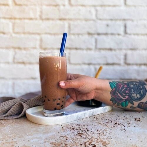 Delicious Chocolate Milk Tea - How To Make Cocoa Boba Recipe 5 glass of chocolate milk tea on a marble tray with a glass of boba, towel on the side, hand reaching for the drink, on a marble and brick background