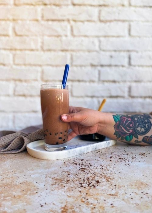 Delicious Chocolate Milk Tea - How To Make Cocoa Boba Recipe 3 glass of chocolate milk tea on a marble tray with a glass of boba, towel on the side, hand reaching for the drink, on a marble and brick background