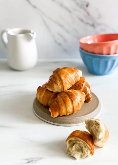 A halved croissant, with its golden crust and soft layers, is the focus, while a plate of croissants in the background adds depth. The soft lighting highlights the crispiness and sheen of the pastry.