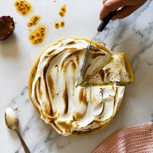 Delicious Passion Fruit Meringue Pie 5 passion fruit meringue pie with a cut slice, hand cutting another slice, on a marble background, passion fruit and seeds on the side, spoon with meringue on the side