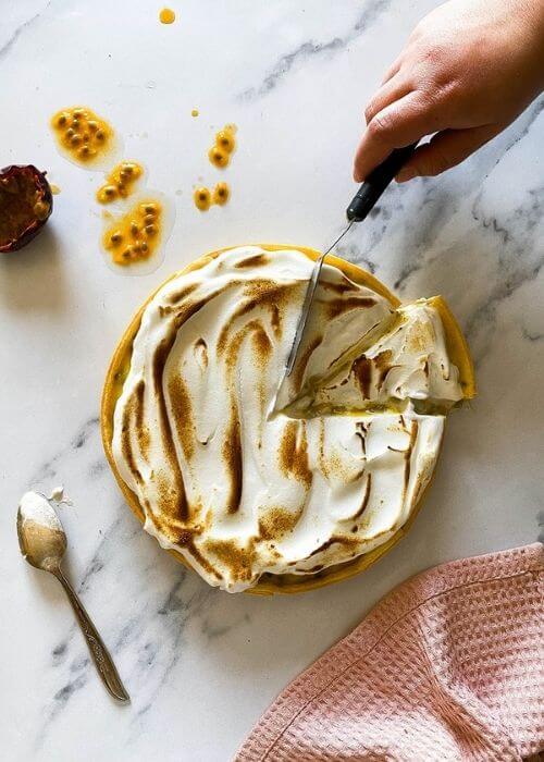 passion fruit meringue pie with a cut slice, hand cutting another slice, on a marble background, passion fruit and seeds on the side, spoon with meringue on the side
