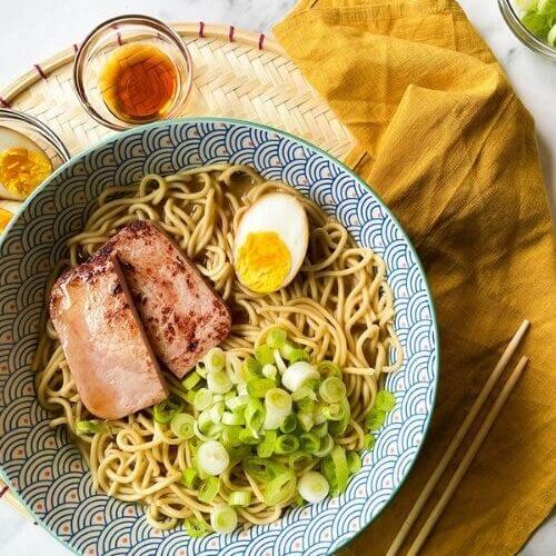 a bowl of ponyo ramen with noodles, spring onions, ham and egg. under a yellow towel, chopsticks, bowls with ingredients on a marble background