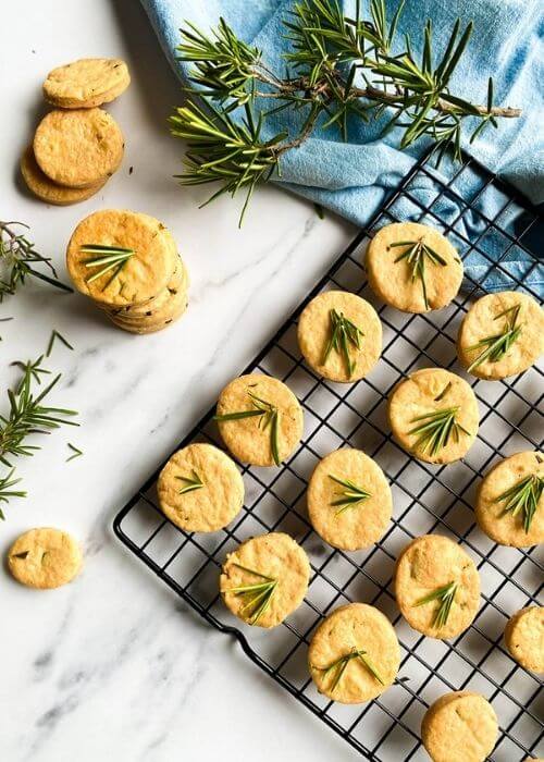Most Delicious Savory Shortbread With Parmesan And Rosemary, 6 savory shortbread cookies with rosemary on top, on a cooling rack, under a blue towel, more cookies around, on a marble background