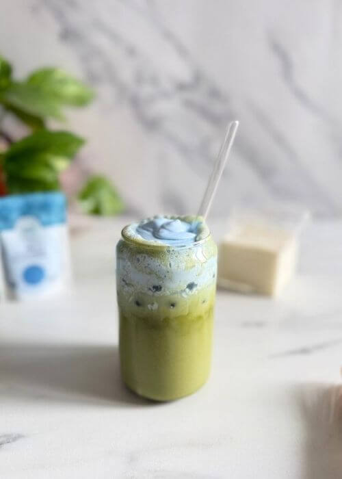 Easy Spirulina Matcha Latte Drink Recipe 6 spirulina matcha latte in a glass with a straw, in the marble background a jug of milk, spirulina powder bag and a plant