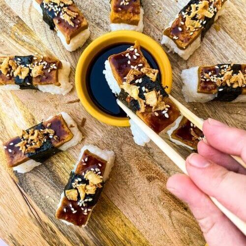Super Delicious Tofu Nigiri With Garlic Soy Sauce 6 hand holding with chopsticks a piece of tofu nigiri, under more tofu nigiri around a bowl of soy sauce, on a wooden board and yellow towel