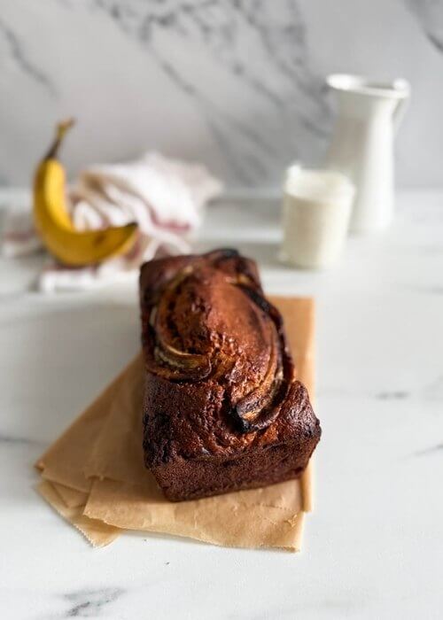 4 ingredient banana bread, on marble background, a glass of milk and towel on the side, banana behind