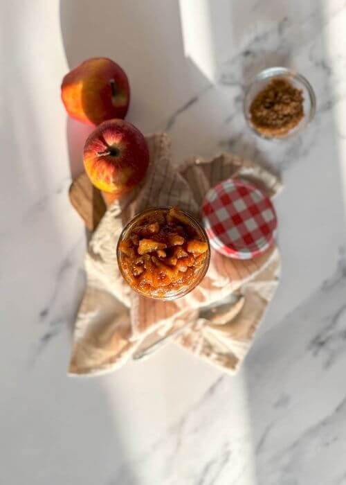 from top - apples caramelized in a jar, over a striped towel, a spoon, apples and brown sugar on the sides, over a marble background