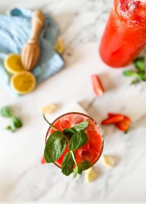 from top - glass of blended strawberry lemonade on a marble coaster, behind a jug of lemonade, strawberries mint and lemons around, in the back a blue towel and lemon squeezer on a marble background