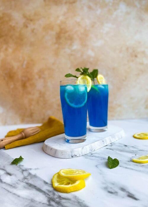 from front - two glasses of blue lagoon fizz chillis recipe on a marble tray and marble background, around a yellow towel, a lemon and mint leaves and lemon squeezer