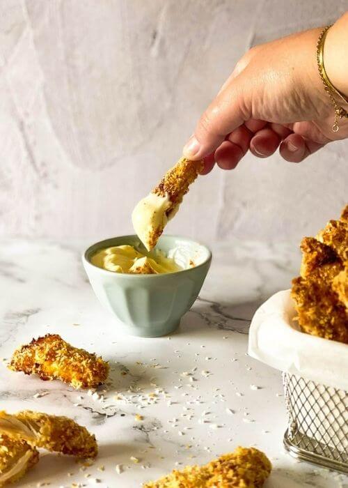 hand dipping one chicken fries in the mayo, chicken fries in a container with parchment paper on a marble background, more fries around and panko, behind a bowl with mayo