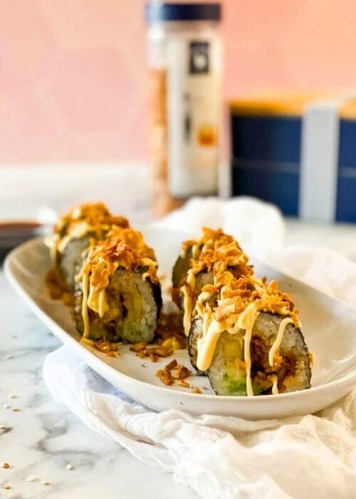 Easy & Quick Chicken Tempura Roll Recipe 7 close up chicken tempura roll on a plate, under a towel, in the background a bento box, crispy onions and chopsticks, on marble table and pink background