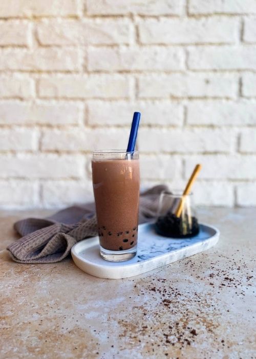 Delicious Chocolate Milk Tea - How To Make Cocoa Boba Recipe 4 glass of chocolate milk tea on a marble tray with a glass of boba, towel on the side, on a marble and brick background