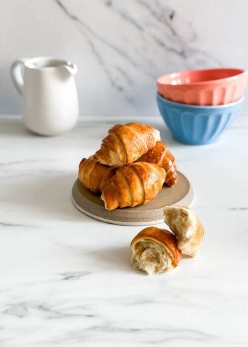 A stack of shiny, air-fried croissants sits on a neutral-toned plate, with a broken croissant in front. In the background, a white milk jug and pastel-colored mixing bowls add a cozy kitchen vibe.