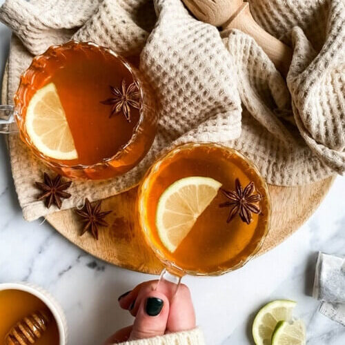 two cups of hot toddy mocktail with slices of lemon and star anise, over a wooden board and light brown tea towel, slices of lemons, star anise and honey around, hand holding one cup