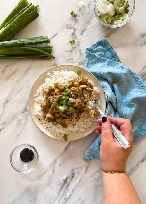 Miso Chicken Aubergine Recipe 2 hand with a spoon miso chicken with aubergine on a plate of rice, under a towel, green onions and garlic seeds in bowls, on a marble background