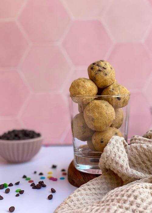The Best No Bake Edible Cookie Dough Bites 3 cookie dough bits in a clear glass, towel and choc chip in a bowl in the back, pink background