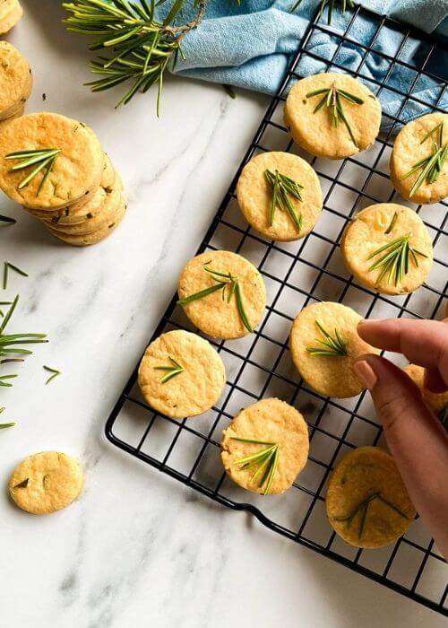 Most Delicious Savory Shortbread With Parmesan And Rosemary, 5 savory shortbread cookies with rosemary on top, on a cooling rack, under a blue towel, more cookies around, on a marble background, hand reaching for one