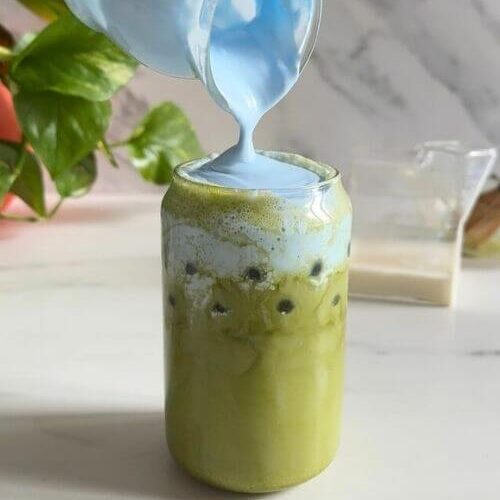 Easy Spirulina Matcha Latte Drink Recipe 8 spirulina matcha latte being made, spirulina cold foam being poured, a plant, a milk jug and chansen in the background, on a marble background