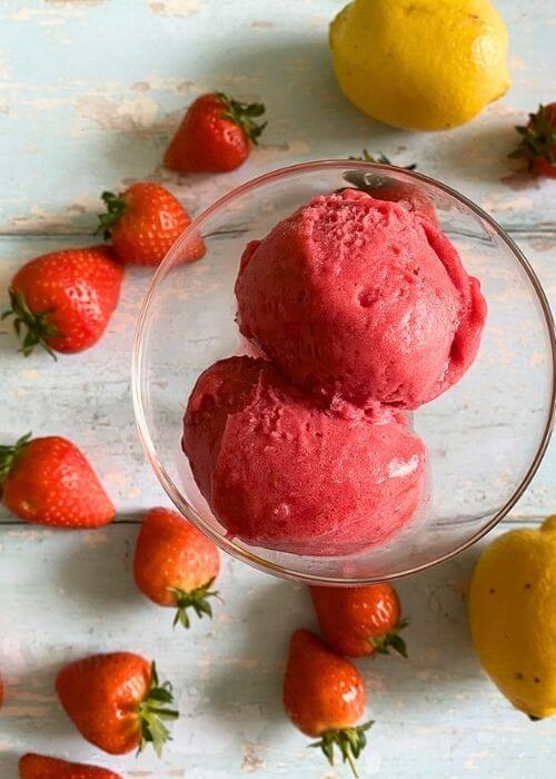 3 Ingredient Strawberry Sorbet Without Ice Cream Maker 1 from top - strawberry sorbet without ice cream maker in a glass bowl, strawberries and lemons around on a light blue background