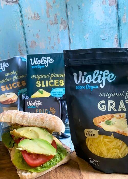 Violife Cheese Review From A Non-Vegan 4 four packs of violife cheese on a wooden board, beside a cheese sandwich, all on a wooden board, blue background