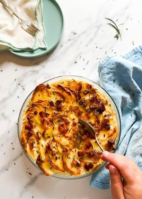 air fryer scalloped potatoes in a tray, under a blue towel, on the side a plate with a towel and cutlery, under a marble background, hand with a spoon taking some potatoes from the tray