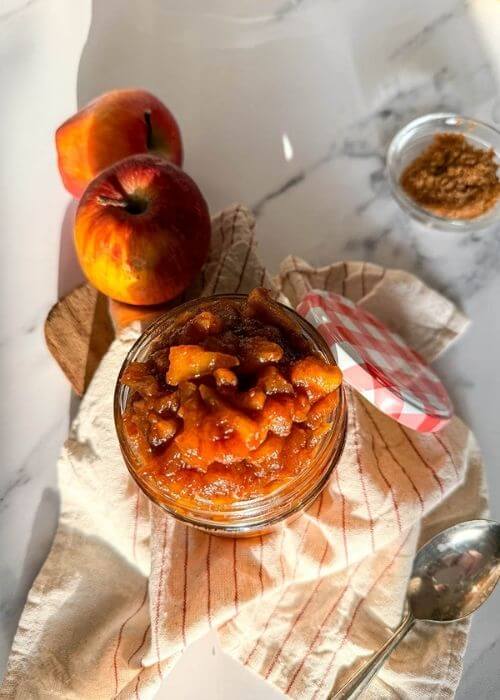 close up apples caramelized in a jar, over a striped towel, a spoon, apples and brown sugar on the sides, over a marble background
