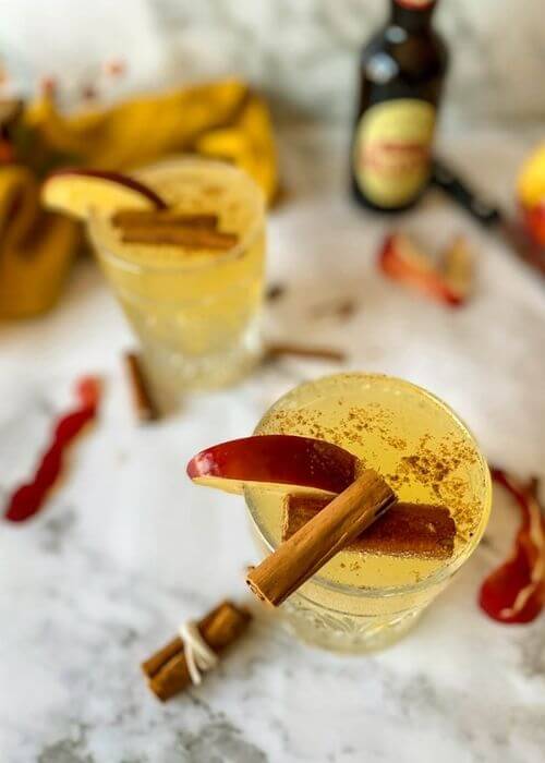 Quick Apple Fizz Mocktail, Delicious Fall Drink 3 close up, two glasses of apple fizz mocktail with apple slices and cinnamon sticks on top, apple peels, cinnamon sticks, knife, towel and leaves around, on marble background, bottle of ginger beer