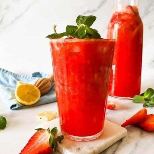 glass of blended strawberry lemonade on a marble coaster, behind a jug of lemonade, strawberries mint and lemons around, in the back a blue towel and lemon squeezer on a marble background