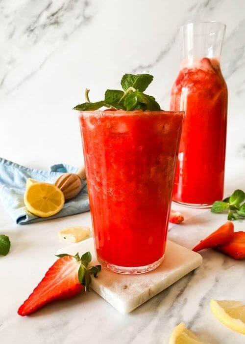 glass of blended strawberry lemonade on a marble coaster, behind a jug of lemonade, strawberries mint and lemons around, in the back a blue towel and lemon squeezer on a marble background