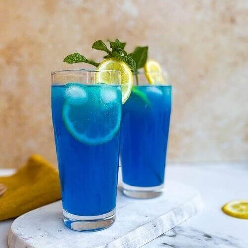 close up - two glasses of blue lagoon fizz chillis recipe on a marble tray and marble background, around a yellow towel, a lemon and mint leaves