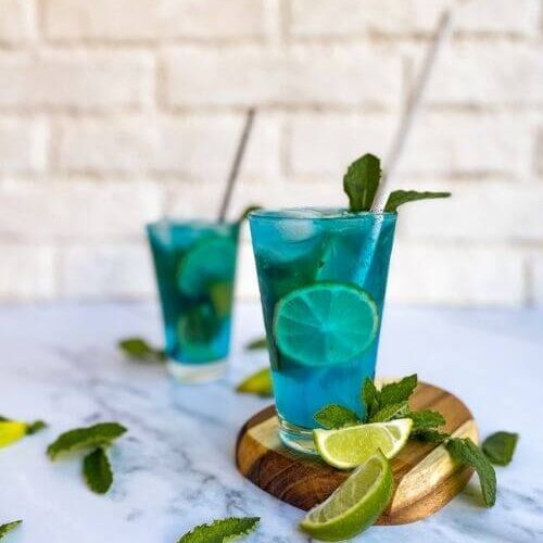 two glasses of blue mojitos on a wooden board, with lime slices and mint leaves on the side. On a white background