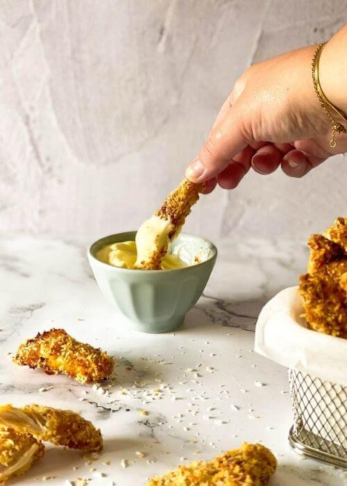 hand dipping one chicken fries in the mayo, chicken fries in a container with parchment paper on a marble background, more fries around and panko, behind a bowl with mayo