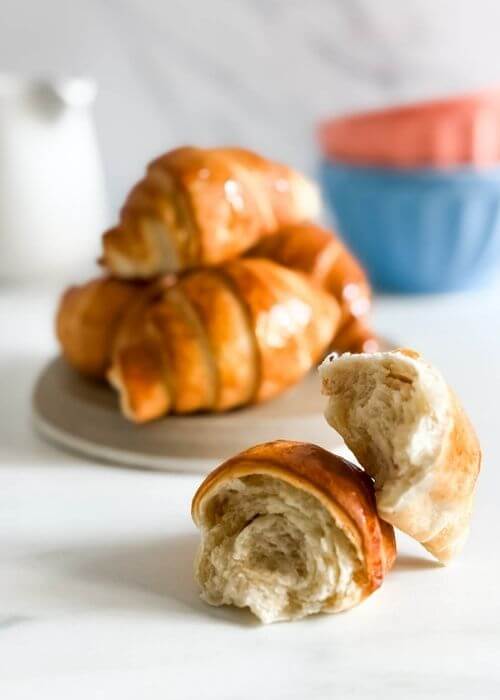 A broken croissant air fryer sits in the foreground, showing off its fluffy, layered interior, while a plate of whole, golden-brown croissants rests behind it on a beige plate.