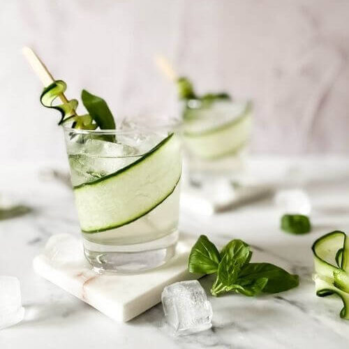 two glasses of cucumber basil mocktail on marble coasters and on a marble background, around ice, basil, cucumber slices and an ice tongs