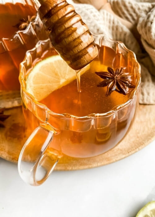 Festive Hot Toddy Mocktail Recipe, Warm & Delicious 5 close up two cups of hot toddy mocktail with honey dripping, with slices of lemon and star anise, over a wooden board and light brown tea towel, slices of lemons, star anise and honey around, hand holding one cup