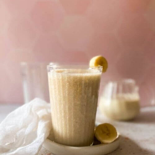 close up banana milk in a glass, on a tray with sliced banana, a towel, another glass and jug behind, a pink and speckled background