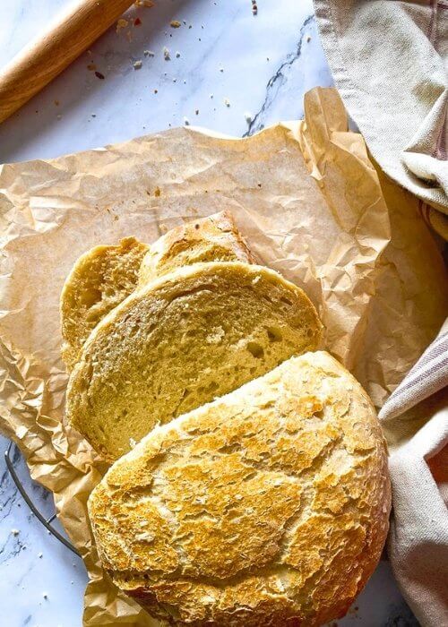 3 Minutes No Knead Bread Without Dutch Oven 5 no knead bread, under parchment paper, wire rack, towel and on the side a bread a dough whisk, on marble background, slices on the paper