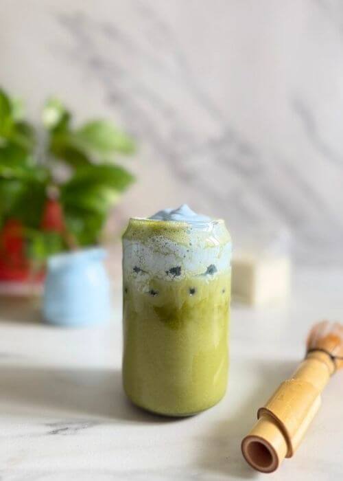 Easy Spirulina Matcha Latte Drink Recipe 7 from front, spirulina matcha latte in a glass, in the marble background a jug of milk, spirulina powder bag and a plant, a chansen