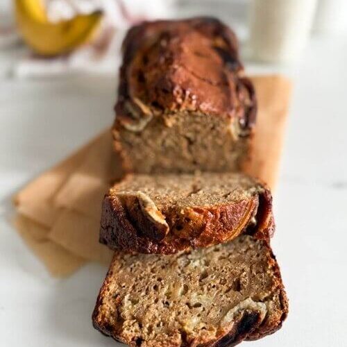 4 ingredient banana bread in a loafpan, on marble background, a glass of milk and towel on the side, banana behind