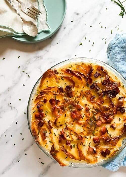 air fryer scalloped potatoes in a tray, under a blue towel, on the side a plate with a towel and cutlery, under a marble background