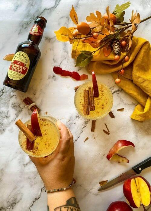 Quick Apple Fizz Mocktail, Delicious Fall Drink 2 from top, one hand holding two glasses of apple fizz mocktail with apple slices and cinnamon sticks on top, apple peels, cinnamon sticks, knife, towel and leaves around, on marble background, glass bottle of ginger beer