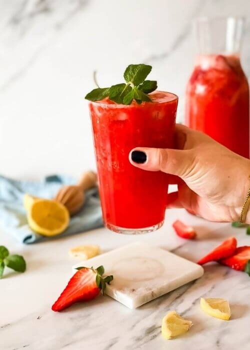 hand holding the. glass of blended strawberry lemonade on a marble coaster, behind a jug of lemonade, strawberries mint and lemons around, in the back a blue towel and lemon squeezer on a marble background