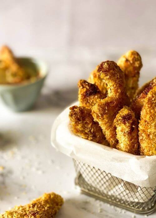close up chicken fries in a container with parchment paper on a marble background, more fries around and panko, behind a bowl with mayo
