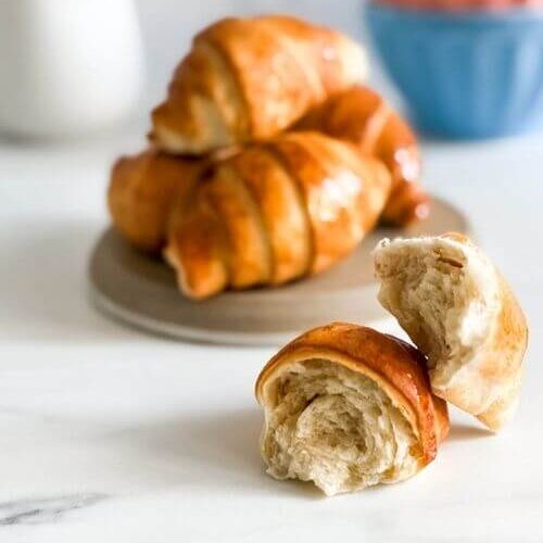 A broken croissant air fryer sits in the foreground, showing off its fluffy, layered interior, while a plate of whole, golden-brown croissants rests behind it on a beige plate.